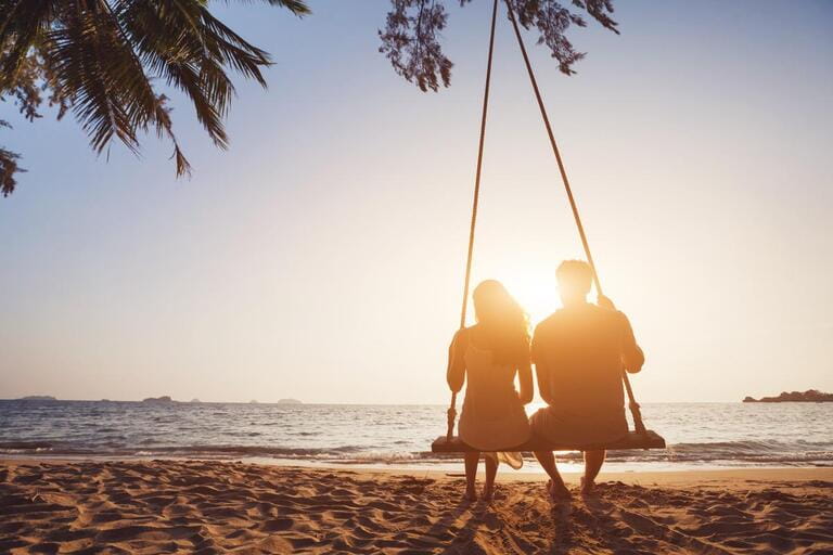 Romantic couple in love sitting together on swing at sunset beach, silhouettes of young man and woman on holidays or honeymoon Romantic couple in love sitting together on swing at sunset beach, silhouettes of young man and woman on holidays or honeymoon