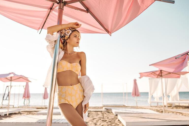 Beautiful caucasian woman is standing on beach crowded with sun loungers with umbrellas. Gorgeous blonde with bandana on her head in yellow swimsuit and white shirt looks to side. Beautiful caucasian woman is standing on beach crowded with sun loungers with umbrellas. Gorgeous blonde with bandana on her head in yellow swimsuit and white shirt looks to side.