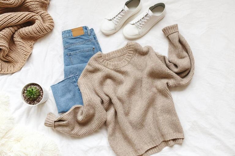 Blue jeans, beige knitted sweater, sneakers, blanket, home plant succulent in a pot lying on bed on white sheet. Overhead view of woman's casual day outfit. Women clothes. Blue jeans, beige knitted sweater, sneakers, blanket, home plant succulent in a pot lying on bed on white sheet. Overhead view of woman's casual day outfit. Women clothes.