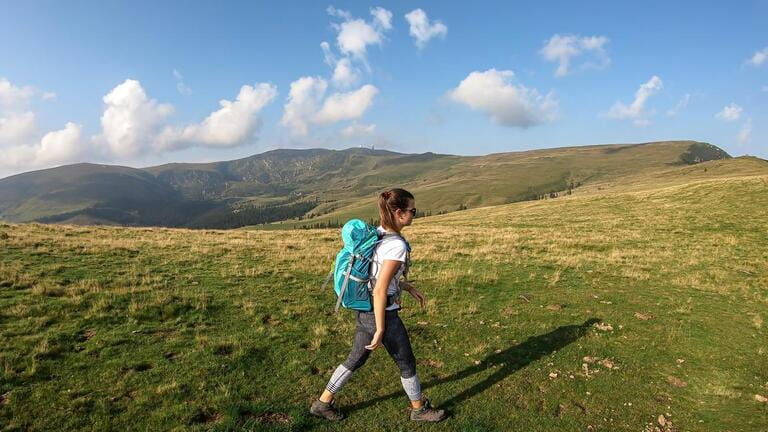 Woman in hiking outfit and big backpack hiking to the top of Speikkogel through a lush pasture in Austrian Alps. The mountains around are green, not so sharp. Sunny summer day. Woman in hiking outfit and big backpack hiking to the top of Speikkogel through a lush pasture in Austrian Alps. The mountains around are green, not so sharp. Sunny summer day.
