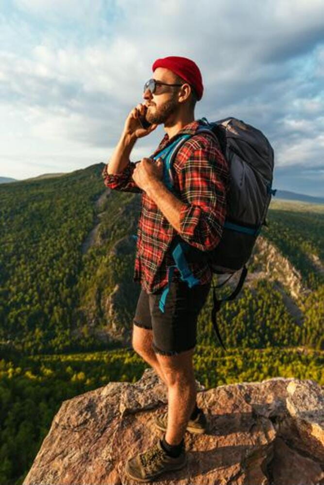 A male traveler in a red hat with a backpack is talking on the phone against the background of mountains. A male traveler in a red hat with a backpack is talking on the phone against the background of mountains.