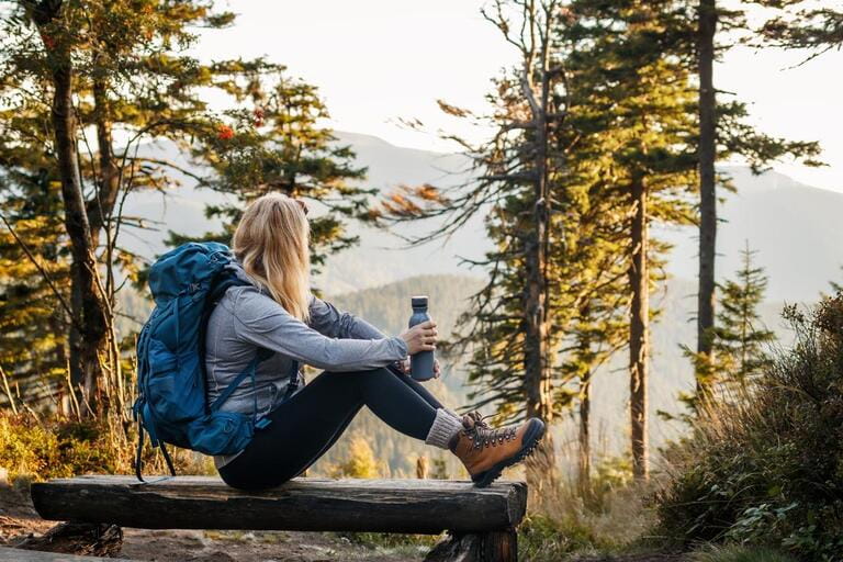 Woman with backpack and thermos resting on bench in forest. Relaxation during hiking in mountain Woman with backpack and thermos resting on bench in forest. Relaxation during hiking in mountain