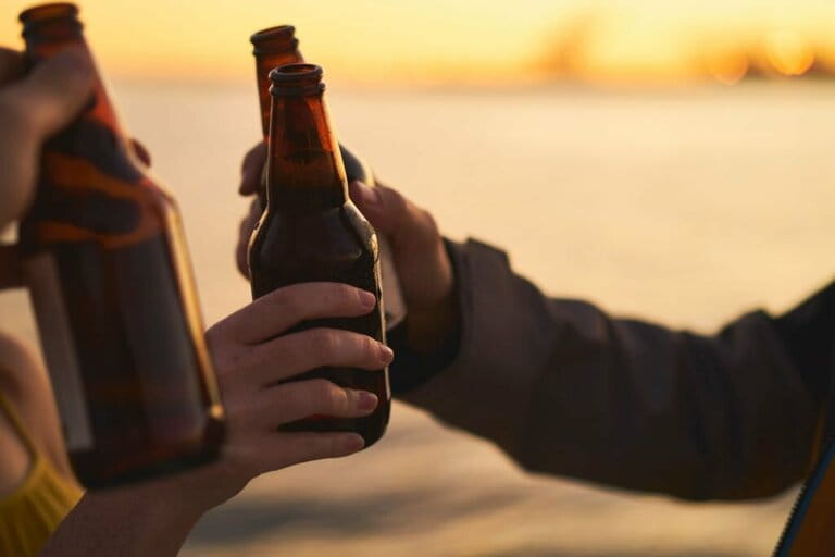 Cropped image of friends toasting beer bottles against sea during sunset Cropped image of friends toasting beer bottles against sea during sunset